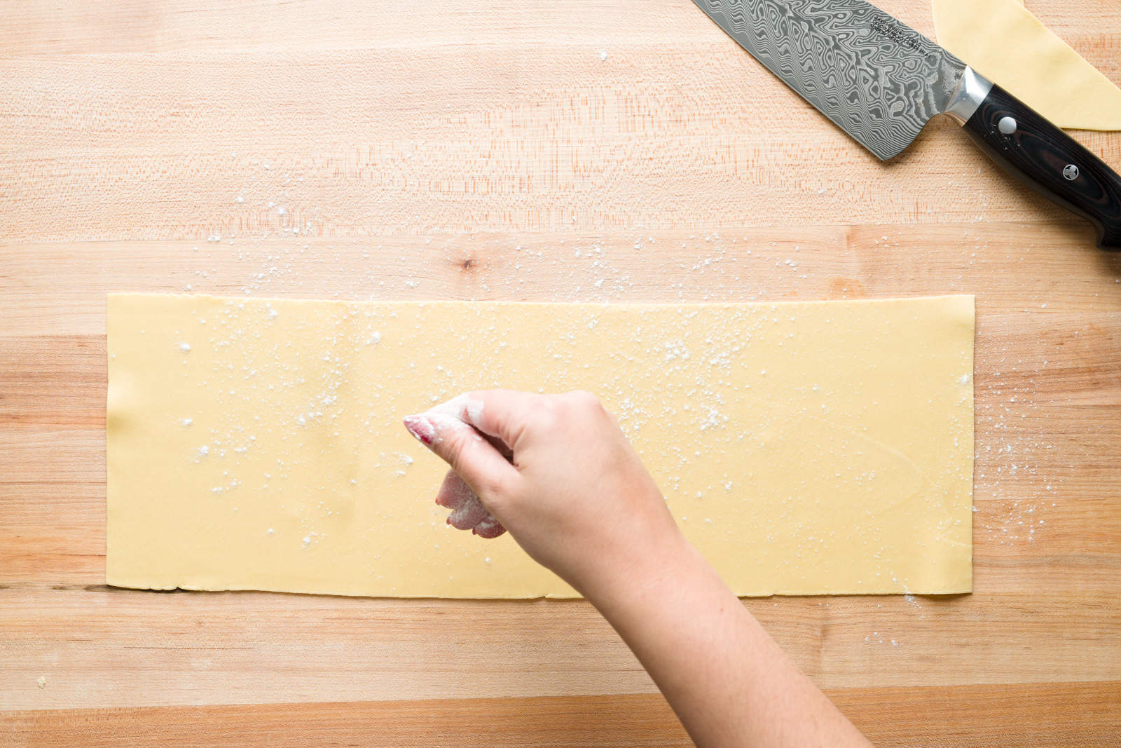 A hand sprinkling flour on a fresh pasta sheet next to a kitchen knife