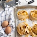 2 ingredient homemade pasta - Fresh pappardelle pasta nests on a baking sheet next to two eggs on a towel next to a pasta machine