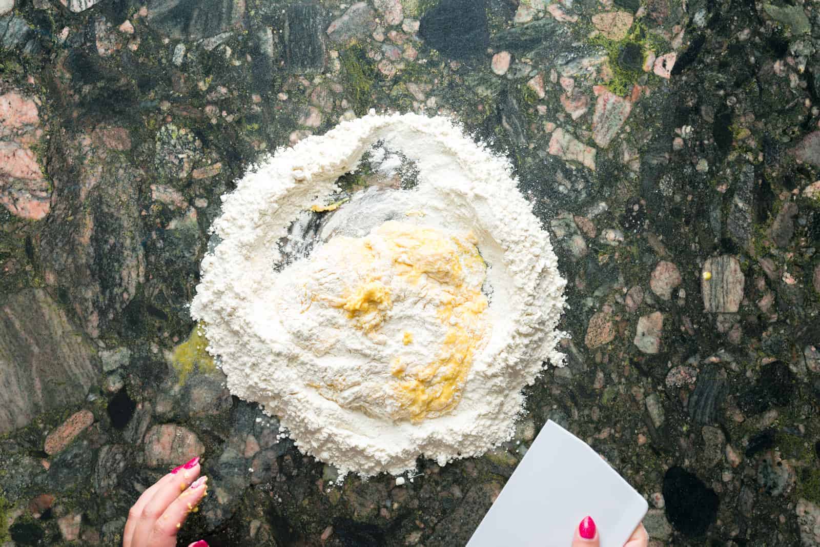 Eggs and flour being scraped together on a kitchen countertop by a person with a dough scraper
