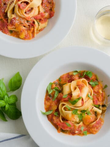 Fresh tomato pasta sauce - Two plates of pasta tossed in fresh tomato sauce in a scene with fresh basil, cherry tomatoes, beverage and cloth napkin