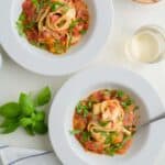 Two plates of pasta tossed in fresh tomato sauce in a scane with fresh basil, cherry tomatoes, beverage and cloth napkin