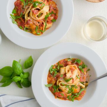 Two plates of pasta tossed in fresh tomato sauce in a scane with fresh basil, cherry tomatoes, beverage and cloth napkin