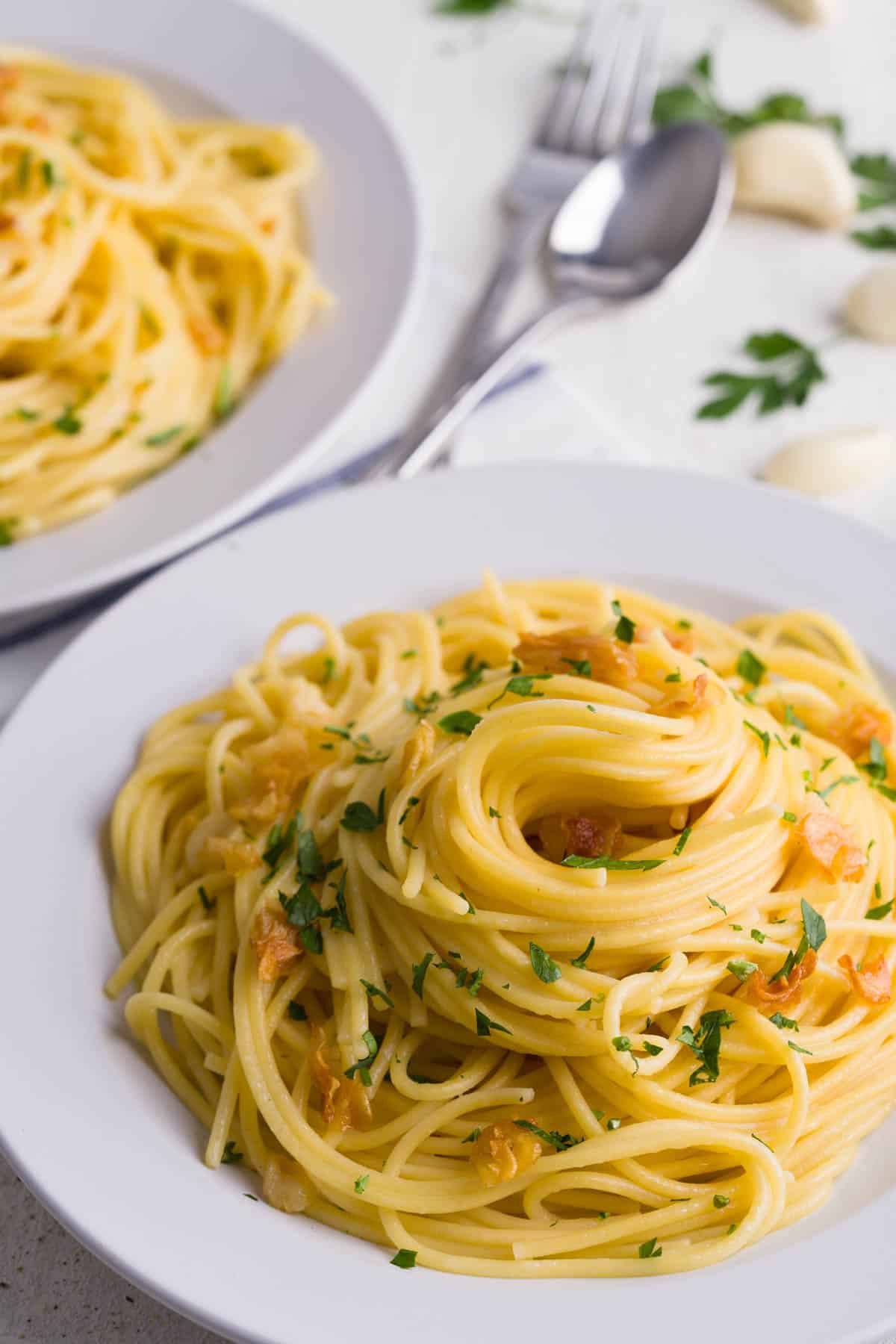 Two plates of spaghetti aglio e olio on a white background around garlic cloves and parsley.