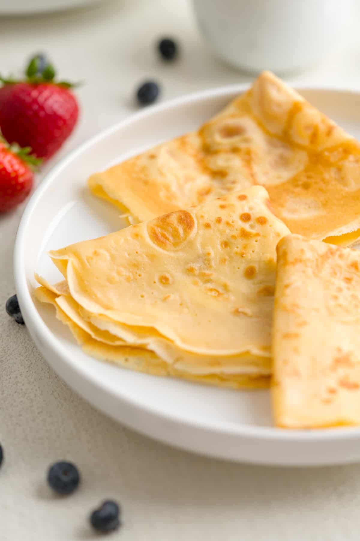 Three simple, folded crepes on a white plate surrounded by strawberries and blueberries.