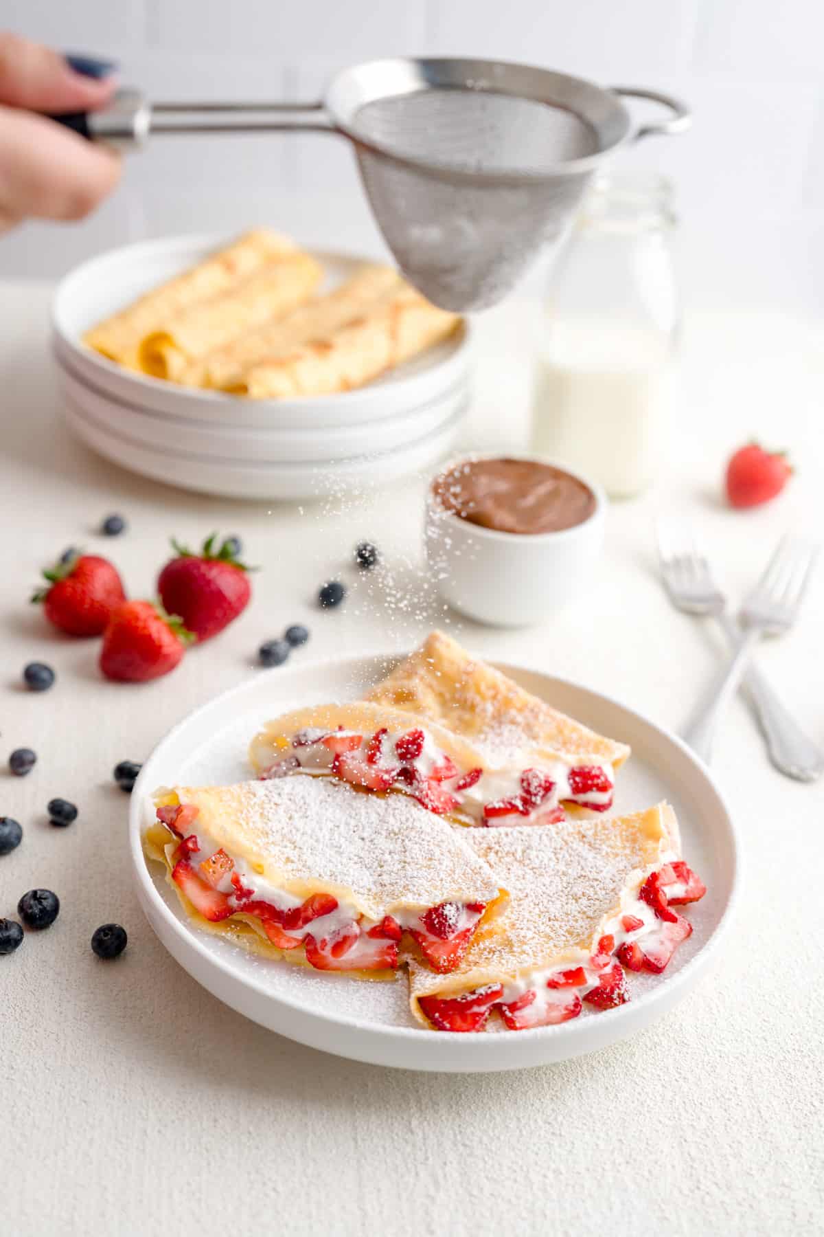 Homemade crepes filled with strawberries and cream dusted with powdered sugar plated in a scene surrounded by blueberries, strawberries and caramel.