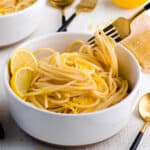 Fork picking up lemon pasta from a white shallow bowl, surrounded by lemons, parmesan cheese and another bowl of lemon pasta