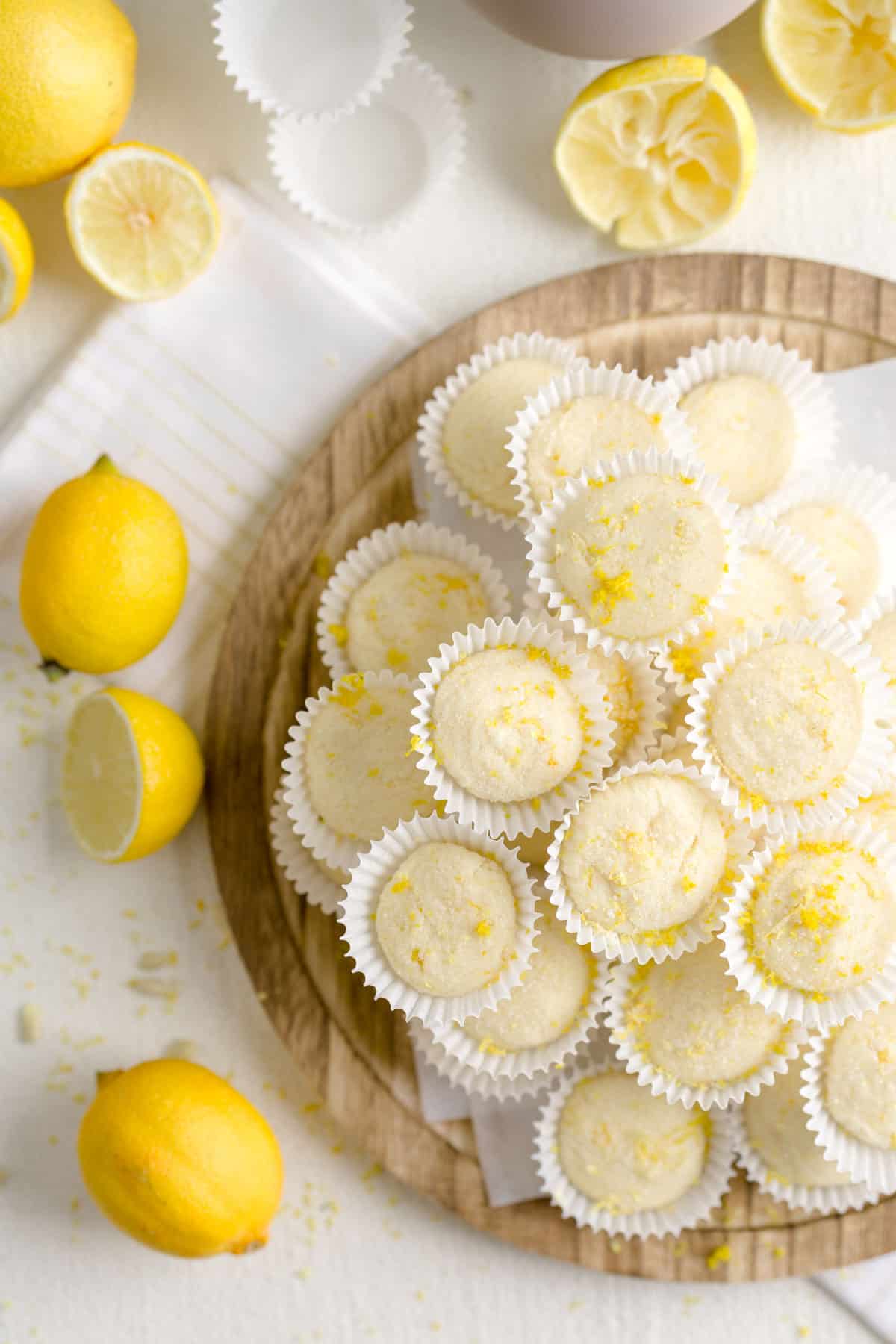 No-bake lemon truffles in mini baking cups stacked on a wooden serving dish surrounded by lemons.