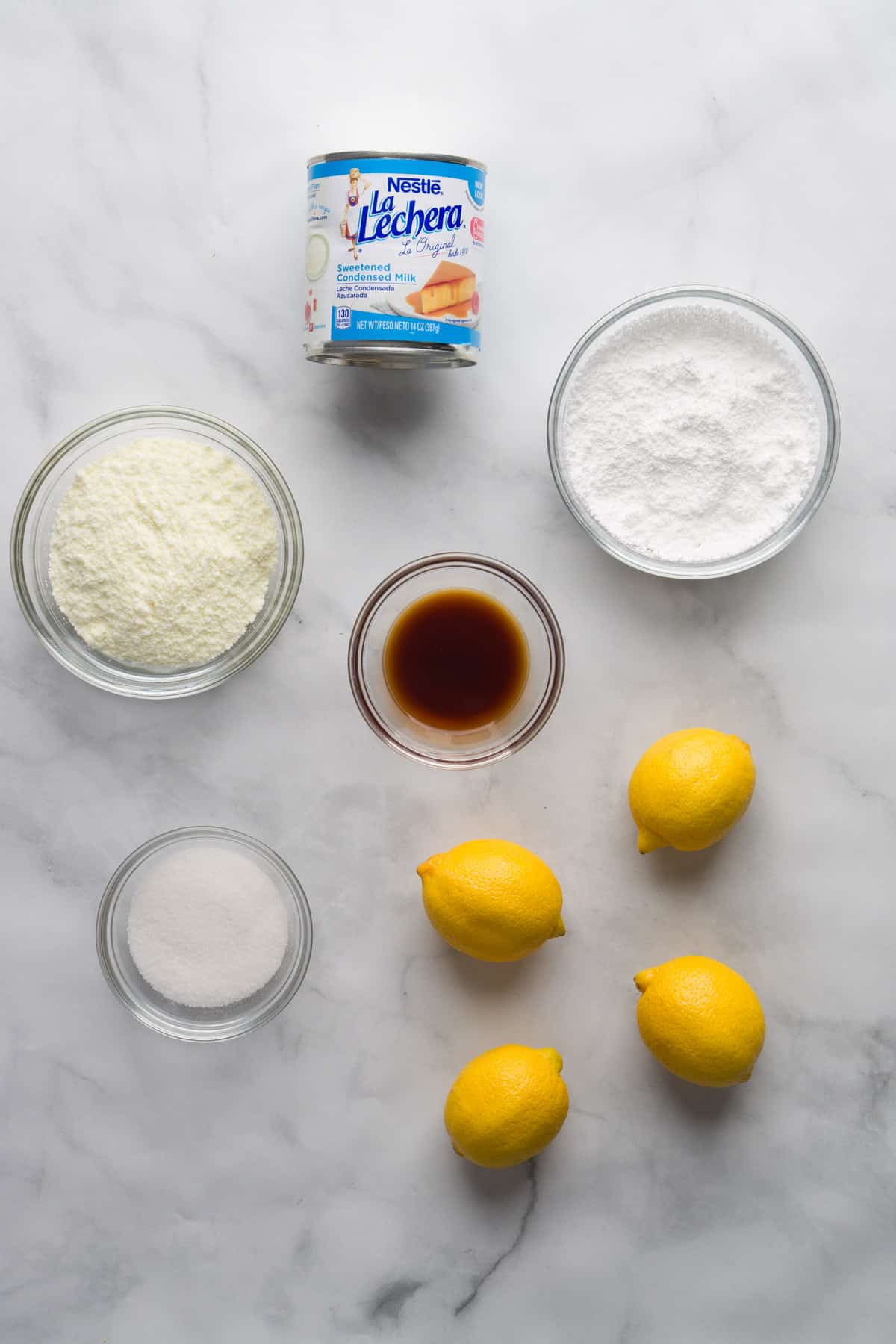Lemon truffle ingredients laid on a marble countertop - lemons, granulated sugar, sweetened condensed milk, powdered milk and powdered sugar.