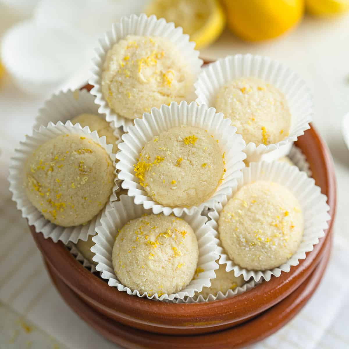 Lemon truffles arranged in white paper cups, topped with lemon zest and placed in a brown ceramic bo