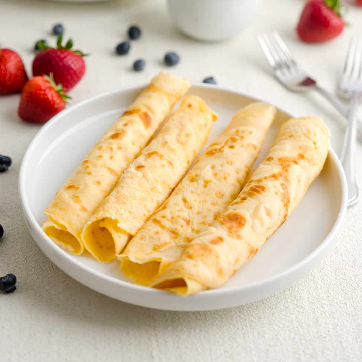 Three golden rolled crepes on a white plate with fresh strawberries, blueberries, and forks in the b