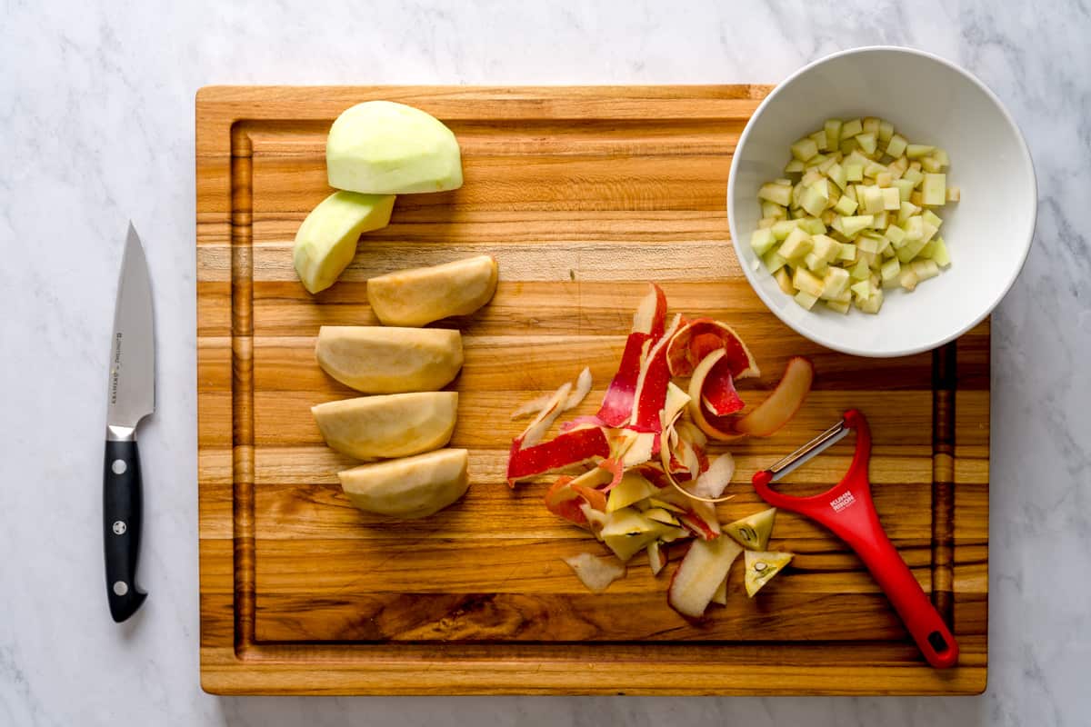 Cut Granny Smith and fuji apples for the apple cake, pictured on a wooden board next to the apple peels and a knife.