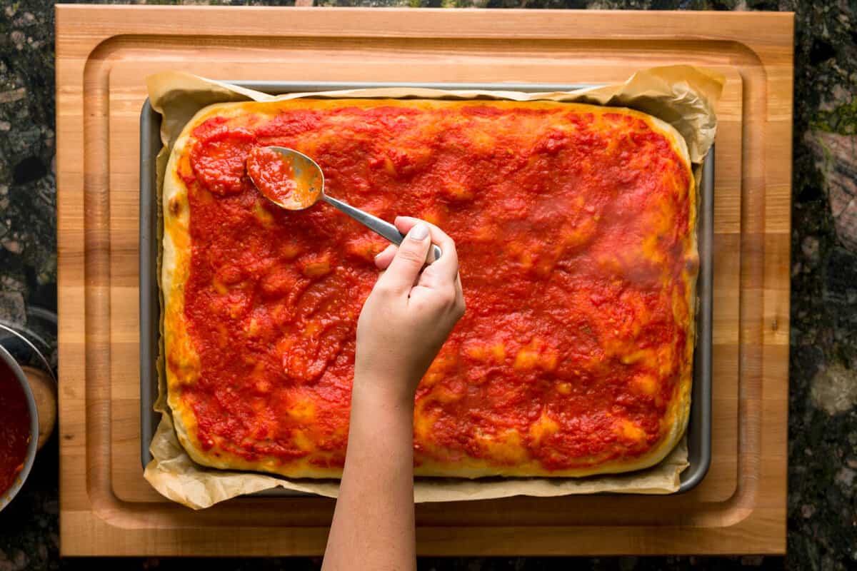 A hand holding a spoon pouring more pizza sauce on a pizza that just came out of the oven.