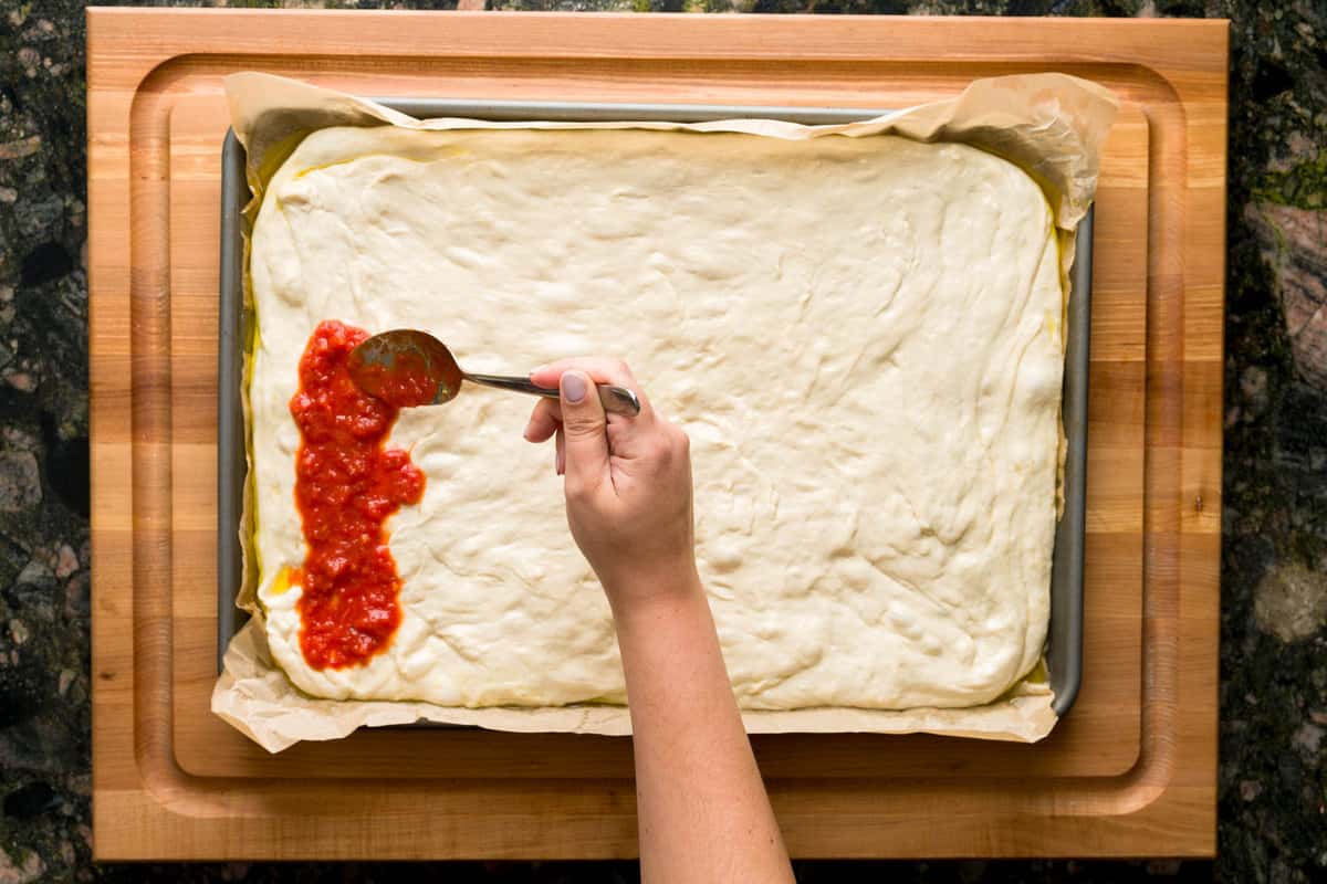A hand spooning pizza sauce on top of stretched Sicilian pizza dough.