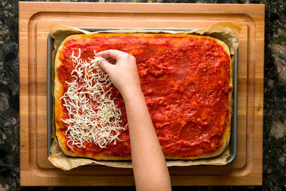 A hand sprinkling mozzarella cheese on a Sicilian sheet pan pizza.