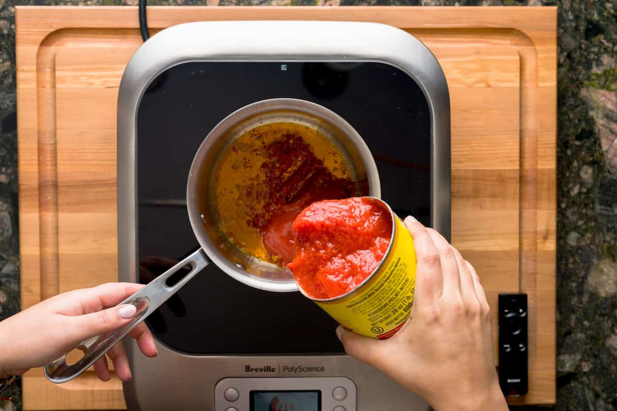 A stove with a pot filled with olive oil, garlic and herbs while a hand pours in whole peeled tomatoes.