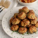 Stack of meatballs on a plate, dusted with parsley, next to a pasta dish and several forks.