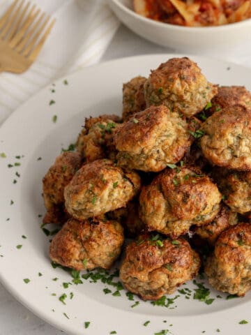 Stack of meatballs on a plate, dusted with parsley, next to a pasta dish and several forks.