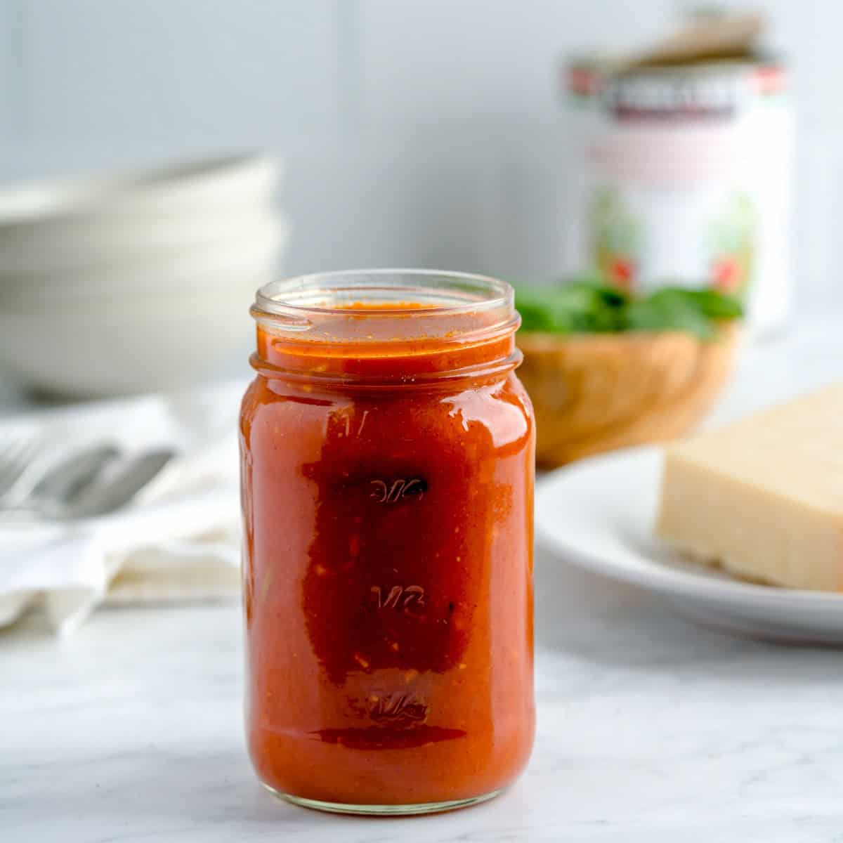 Jar of San Marzano tomato sauce on marble counter with parmesan and herbs in the background.
