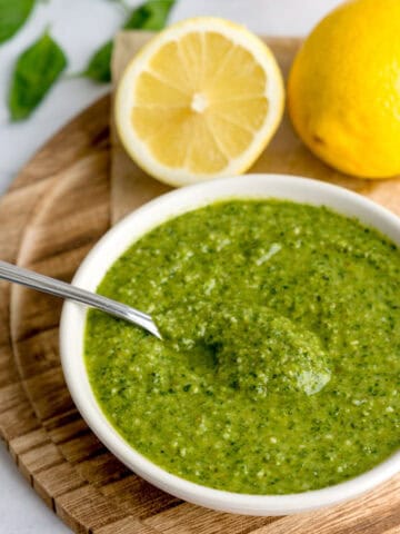 Bowl of easy lemon basil pesto surrounded by a spoon, lemons, basil and walnuts.