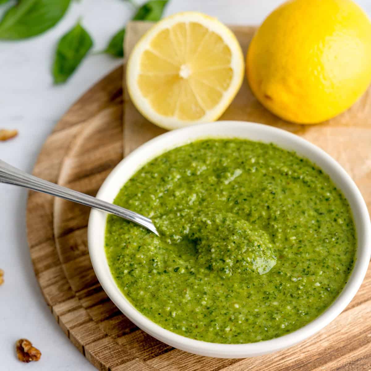 Bowl of vibrant green walnut pesto with a spoon, next to a halved lemon and whole lemon on a wood bo