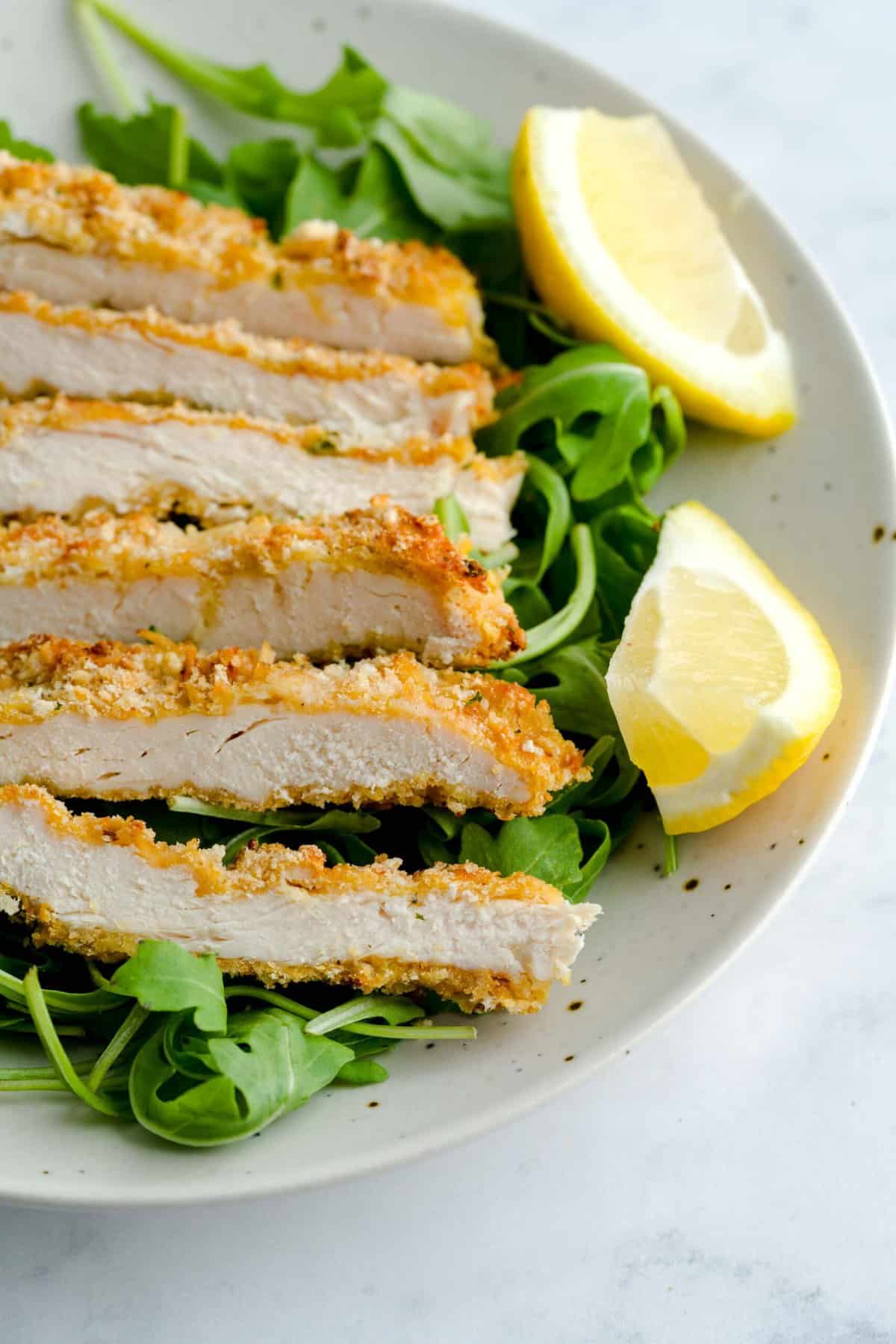 Sliced chicken cutlet on a plate, atop a bed of arugula served with slices of lemon.