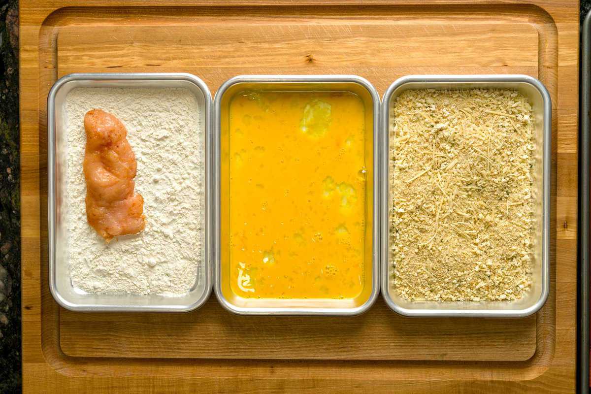 Chicken cutlets breading station, featuring a chicken breast on the tray with all purpose flour, next to a tray with scrambled eggs and seasoned breadcrumbs.