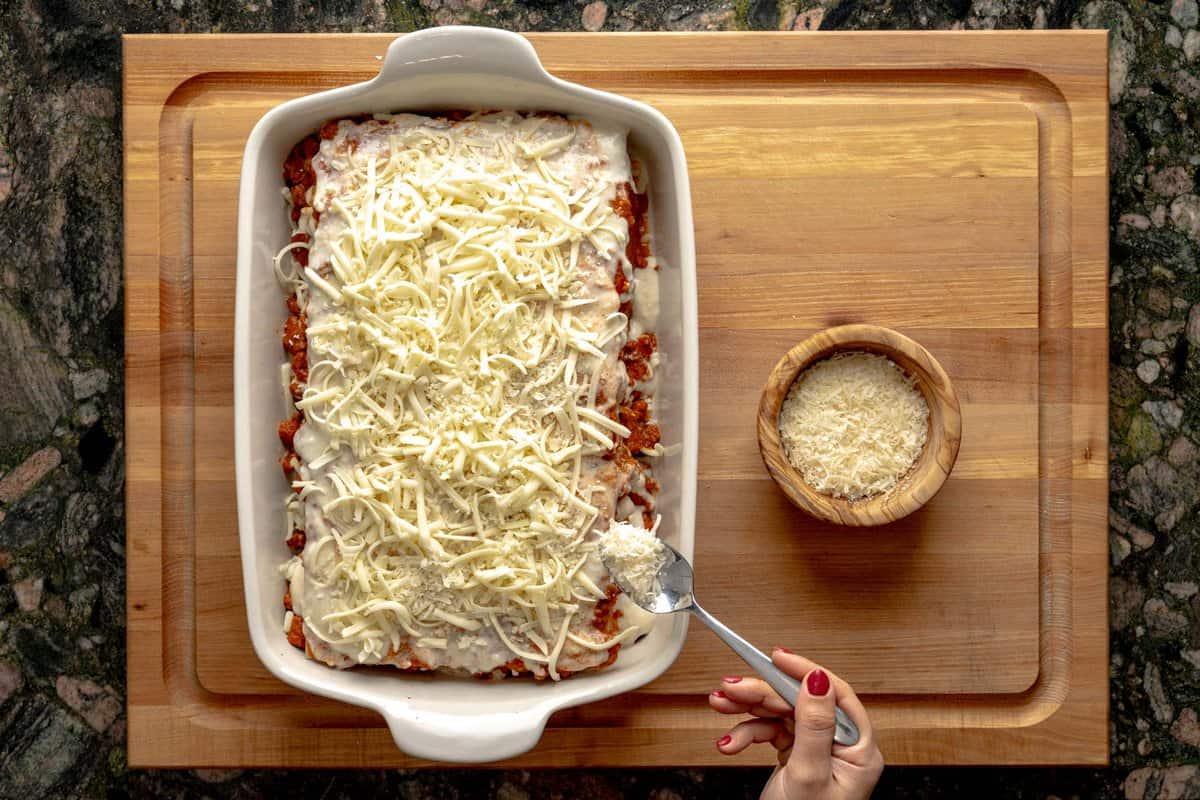 Lasagna being assembled on a lasagna pan, a hand sprinkling parmesan cheese above the lasagna and a bowl with grated parmesan cheese next to it.