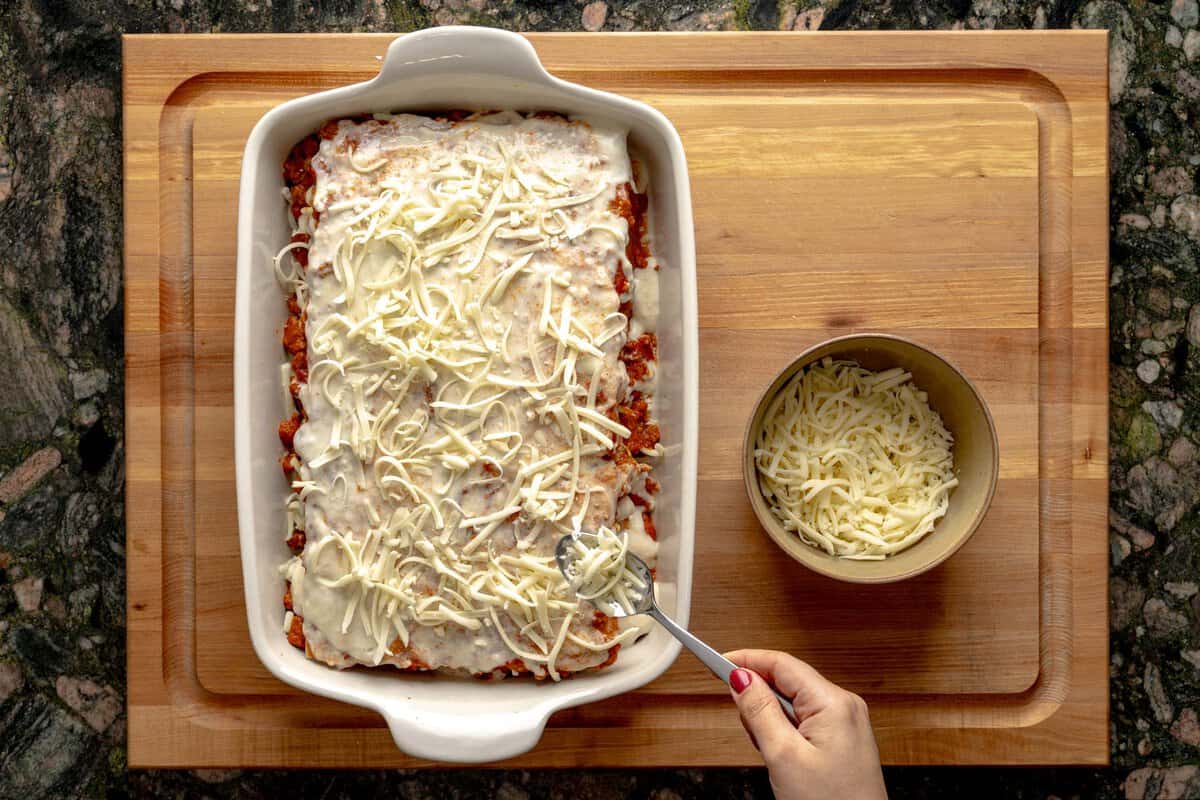 Lasagna being assembled on a lasagna pan, a hand sprinkling mozzarella cheese above the lasagna and a bowl with shredded mozzarella cheese next to it.