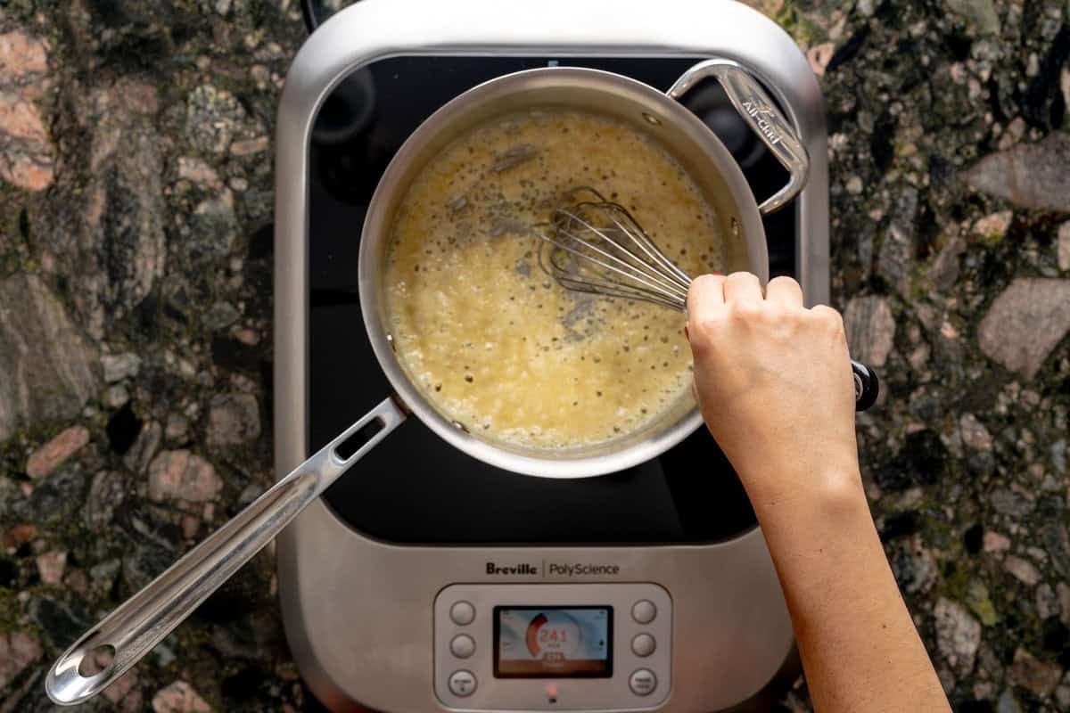 Above shot of pot, above a hot plate, with roux for a shallot béchamel sauce recipe with a hand with a whisk whisking. 
