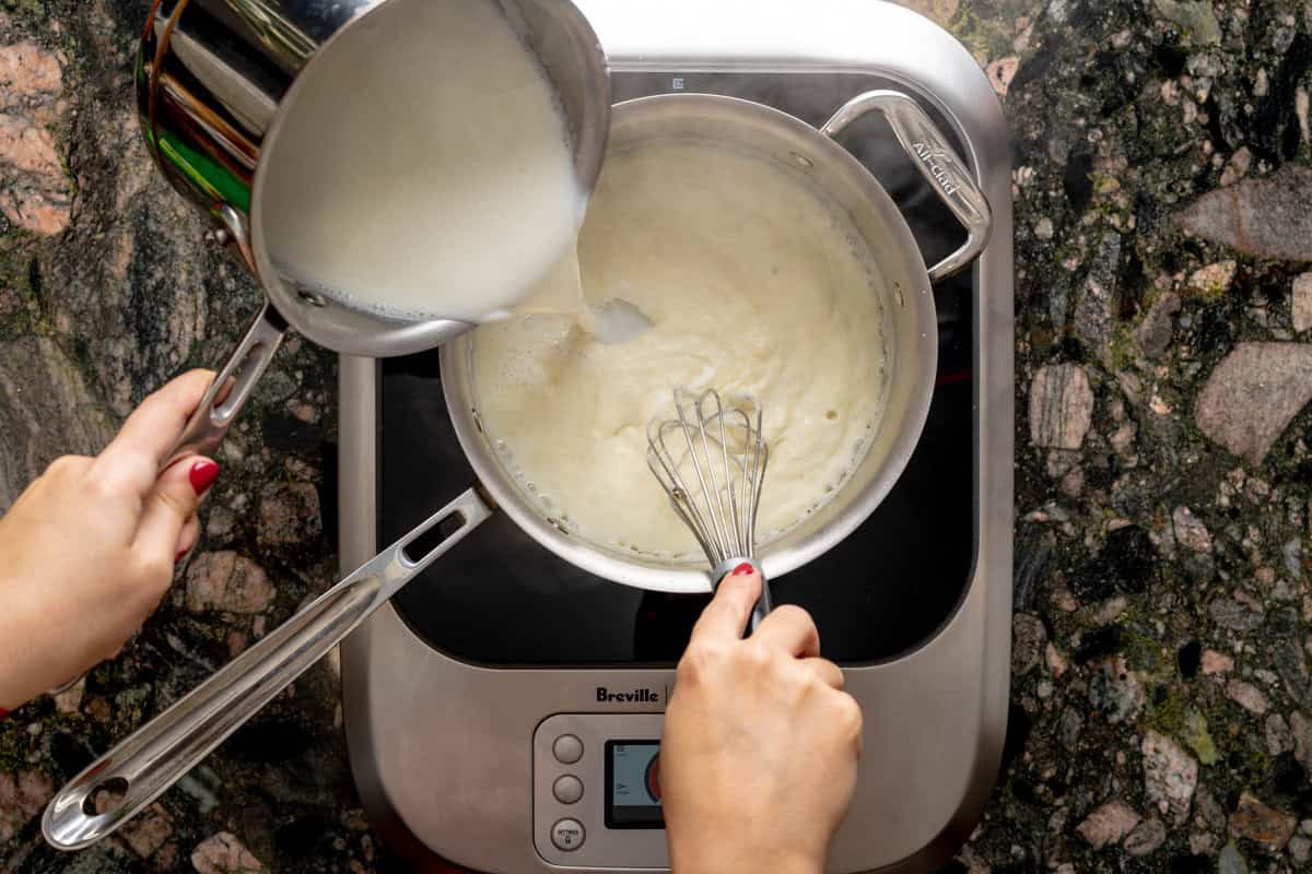 Above shot of pot, above a hot plate, with one hand whisking the roux for béchamel while the other hand pours warm milk into the pot with the roux.