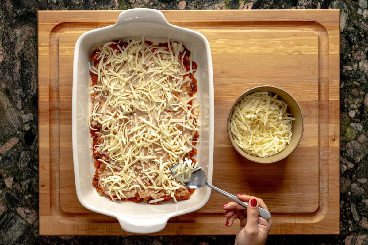 Lasagna being assembled on a lasagna pan, a hand sprinkling mozzarella cheese with a spoon above the lasagna and a bowl with shredded mozzarella cheese next to it.