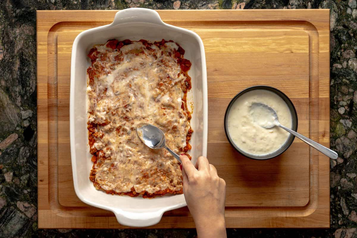 Lasagna being assembled on a lasagna pan, a hand smoothing béchamel sauce with a spoon above the lasagna and a bowl with béchamel sauce next to it.