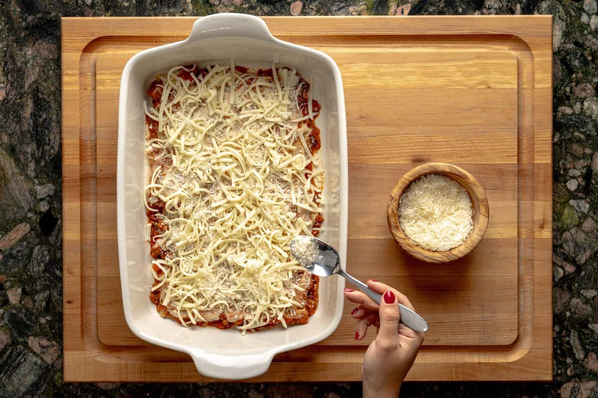 Lasagna being assembled on a lasagna pan, a hand sprinkling parmesan cheese with a spoon above the lasagna and a bowl with grated parmesan cheese next to it.