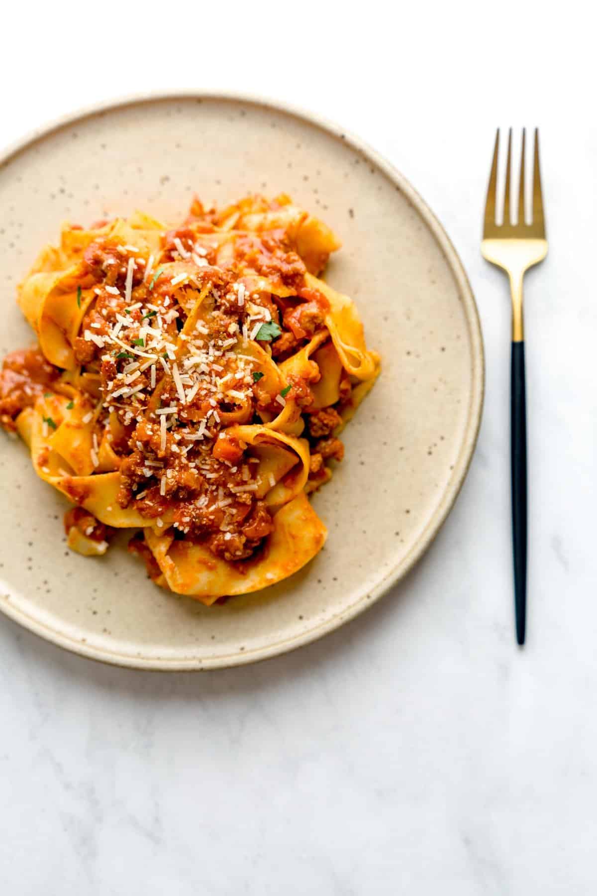 A speckled brown plate with pappardelle bolognese, sprinkled with parmesan cheese and parsley, next to a golden fork.