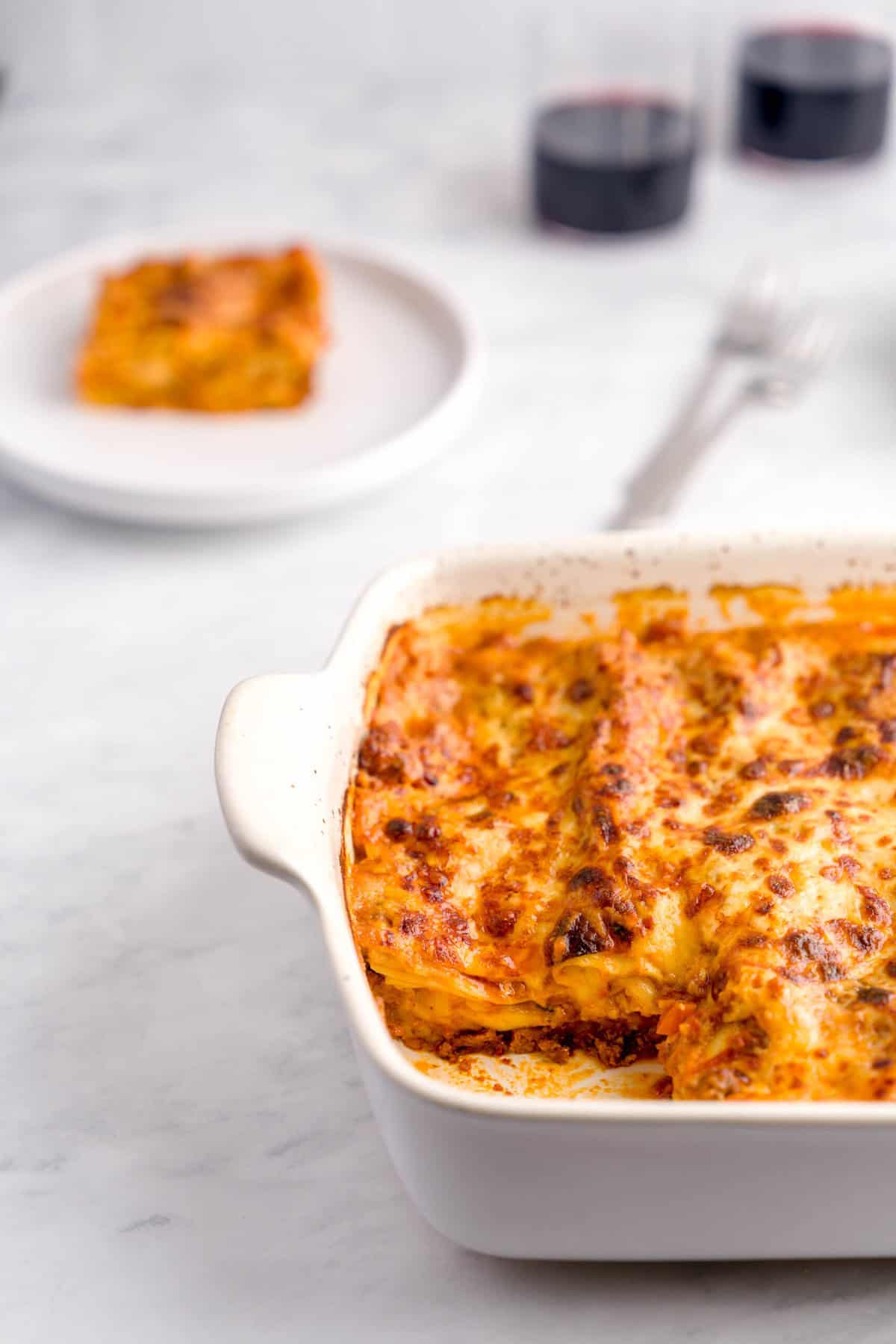 Shot of lasagna in a lasagna pan, with a missing piece which is plated in a pate behind the pan next to two cups of red wine.