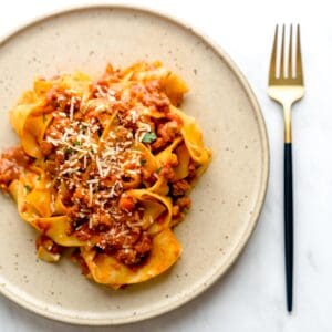 A speckled brown plate with pappardelle bolognese, sprinkled with parmesan cheese and parsley, next to a golden fork.