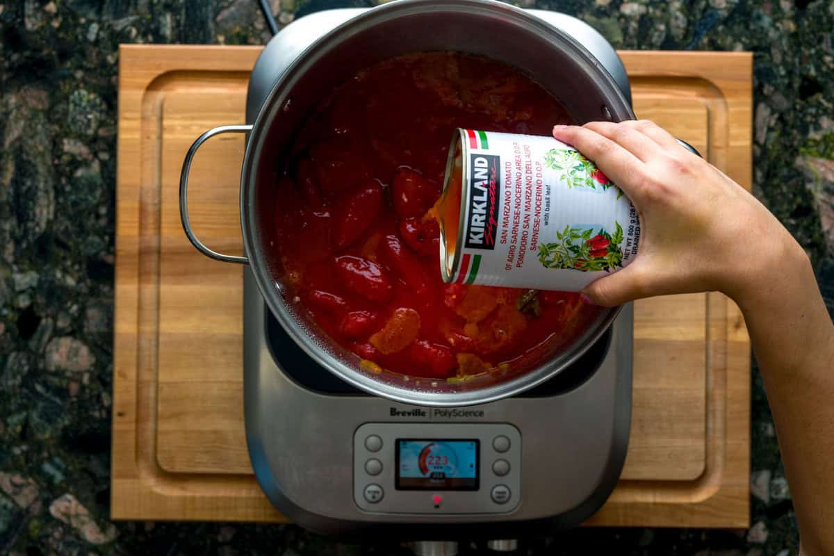 A top-down shot of a large pot over an induction burner. Inside the pot are sautéed veggies, pork and meat and a hand is pouring tomatoes from an open can of whole, peeled tomatoes.