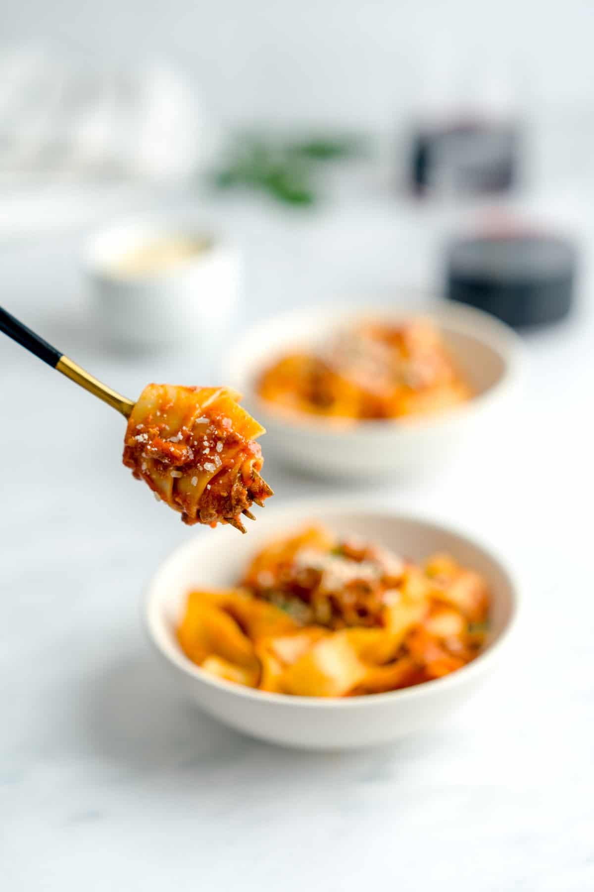 Close up shot of a golden fork holding a twirled tagliatelle with bolognese sauce, sprinkled with parmesan cheese. In the background, two plates of tagliatelle bolognese along with red wine, parmesan cheese and basil in the background.