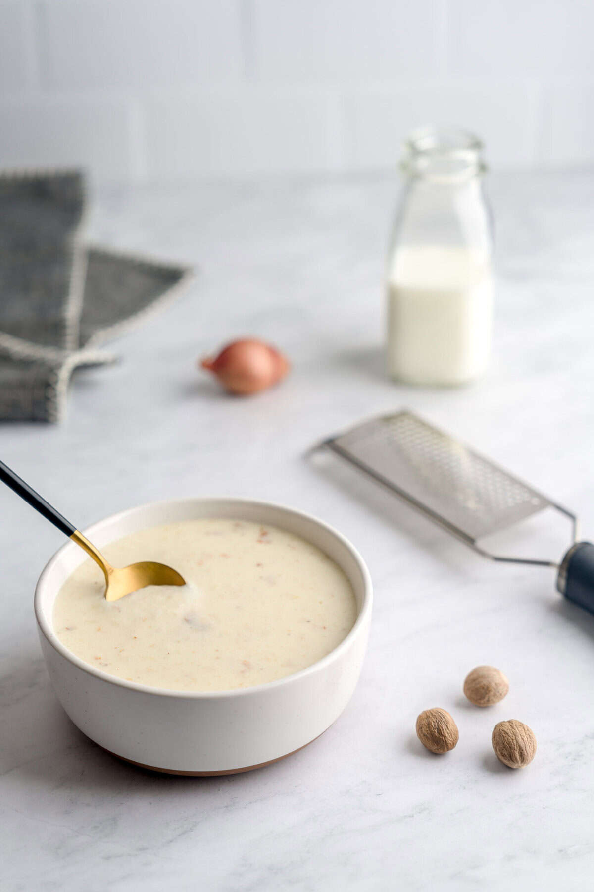 Shot of shallot béchamel sauce in a bowl with a golden spoon dipping into the bowl. Also pictured around the bowl are ingredients for the béchamel, like nutmeg, shallots and milk, along with a gray cloth napkin and a zester.