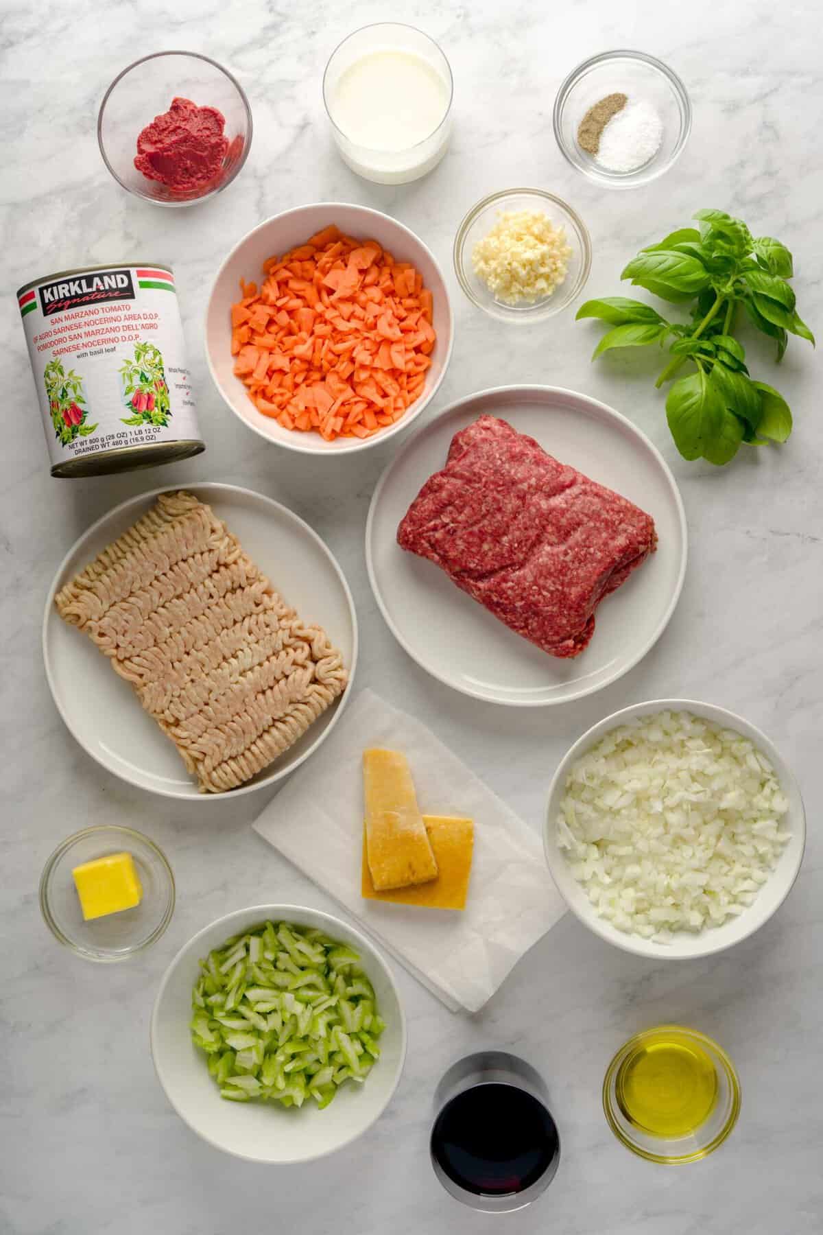 Ingredients for bolognese sauce in an top-down shot, including: ground pork, ground beef, onions, celery, carrots, garlic, milk, red wine, canned, whole tomatoes, tomato paste, salt, pepper, butter, olive oil and basil.