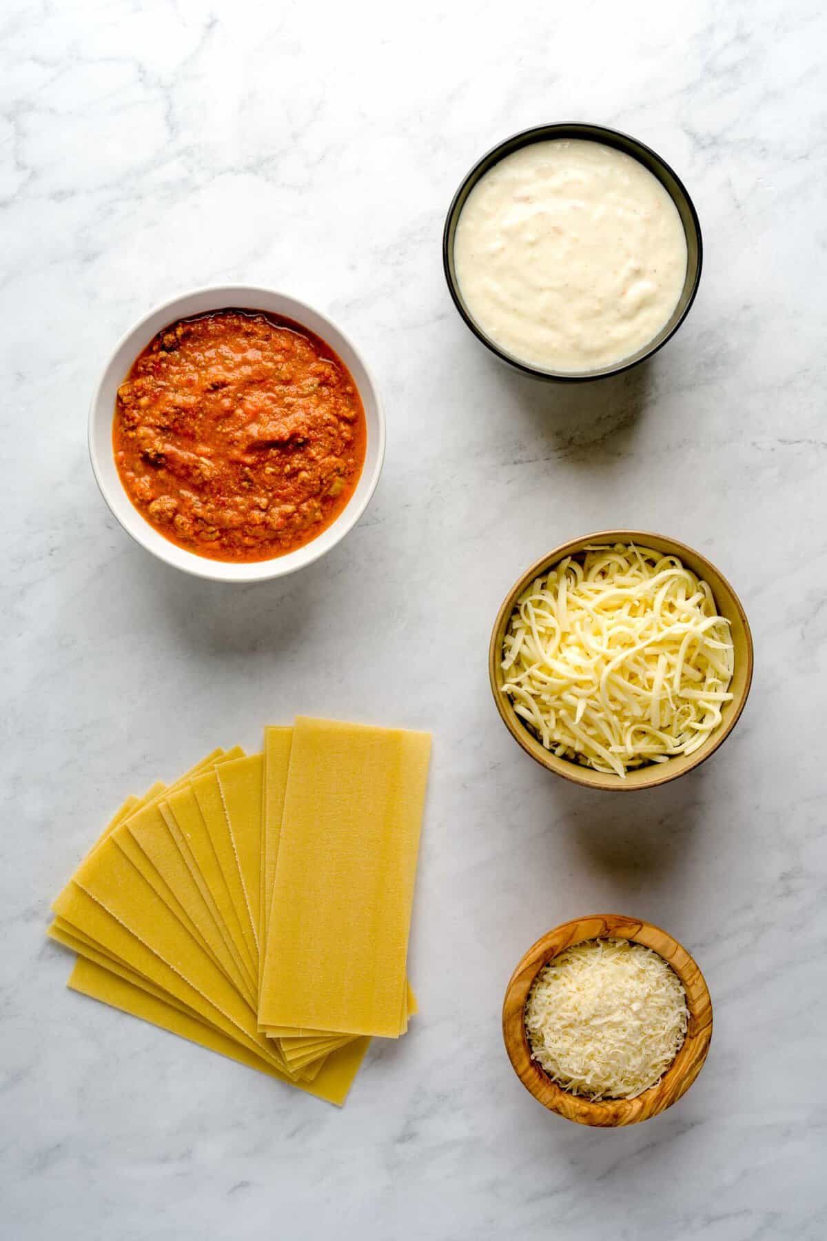 Above shot of the ingredients to make a lasagna bolognese, no-boil lasagna noodles, grated parmesan cheese, shredded mozzarella cheese, shallot béchamel sauce and bolognese sauce, laid out on a marble surface.