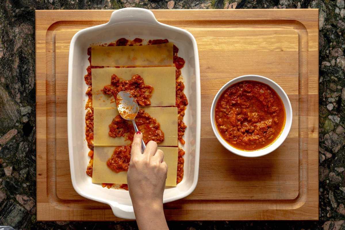 Lasagna being assembled on a lasagna pan, a hand with a spoon pouring béchamel sauce on the lasagna layers with a bowl of béchamel sauce next to the pan.