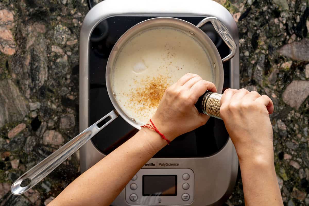 Above shot of pot, above a hot plate, with two hands grinding white peppercorns. In the pot, béchamel sauce is shown along with freshly ground nutmeg and salt at the top of the sauce.