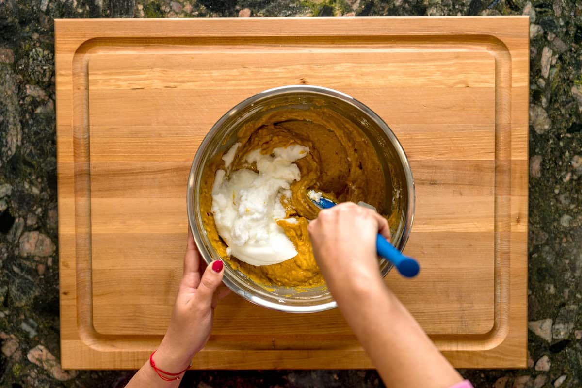 A hand folding the whipped egg whites into the cake batter of the pumpkin spice cake in a metal bowl.