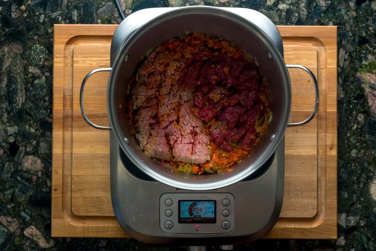 A top-down shot of a large pot over an induction burner. Inside the pot are sautéed carrots, onions and celery along with ground pork and ground meat.