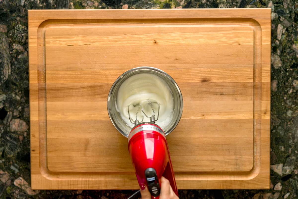 A hand using an electric handheld mixer beating egg whites in a metal bowl.