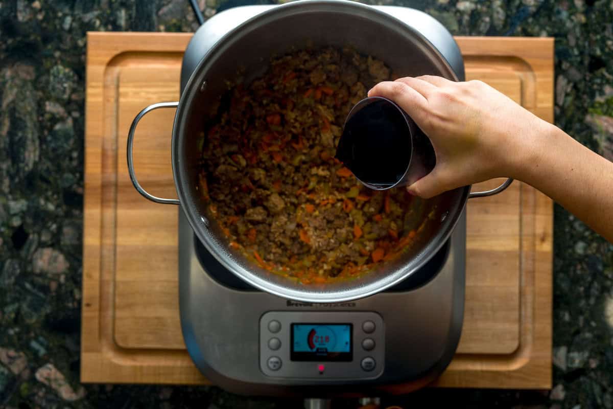 A top-down shot of a large pot over an induction burner. Inside the pot are sautéed veggies, pork and meat and a hand is pouring a glass of red wine into the pot.