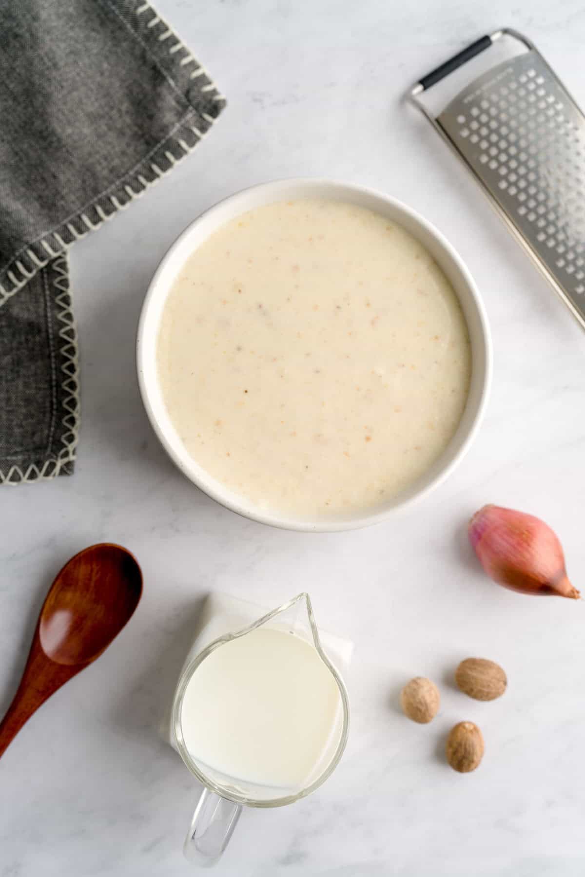 Overhead shot showing shallot béchamel in a white bowl surrounded by a gray cloth napkin, zester, shallot, nutmegs, milk and wooden spoon.