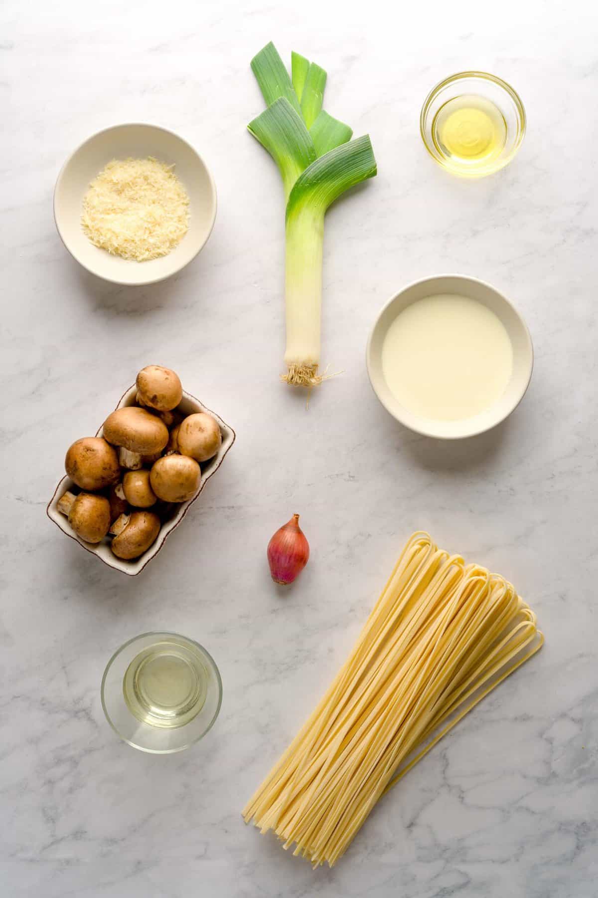 Ingredients laid out for creamy leek and mushroom pasta: white wine, pasta, shallot, leek, baby Bella mushrooms, heavy cream, extra virgin olive oil and grated parmesan cheese.