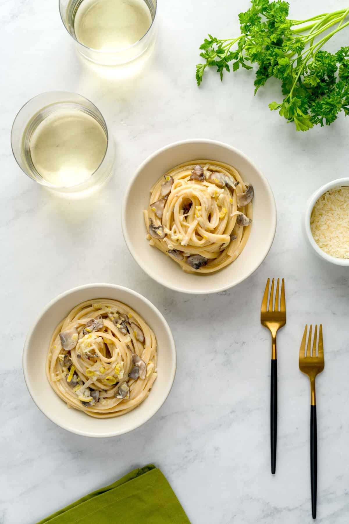 Two bowls of creamy leek and mushroom pasta staged on a marble countertop next to a green napkin, two forks, grated parmesan cheese, white wine glasses and parsley.